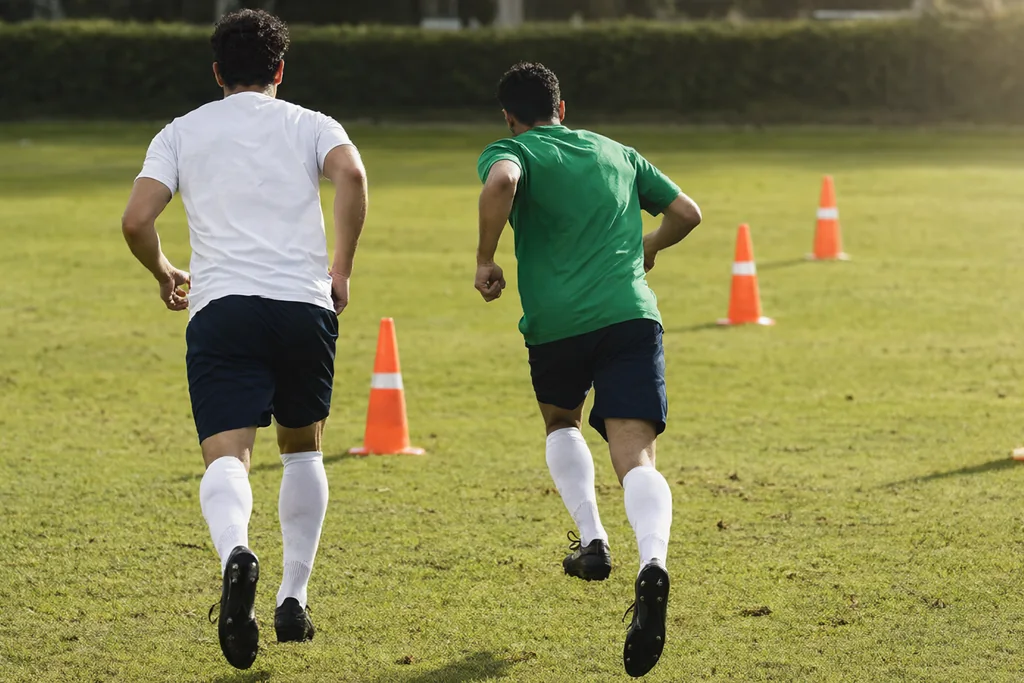 Dois jogadores de uniforme liso em treino de passe em campo verde, vistos de costas