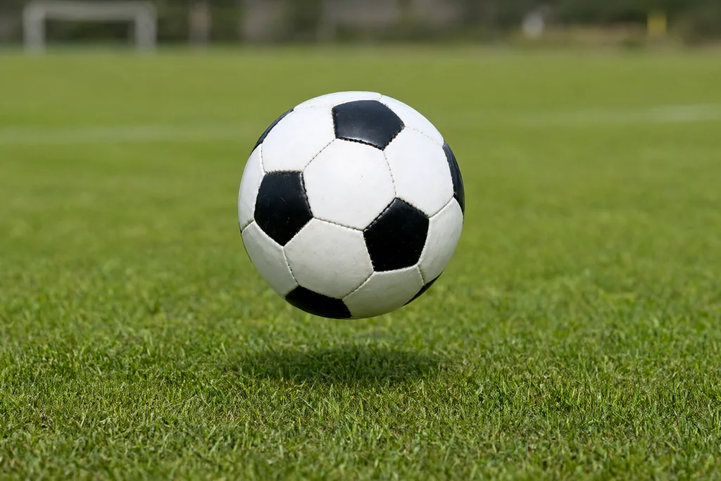 Bola de futebol redonda no ar sobre gramado verde com jogadores desfocados ao fundo